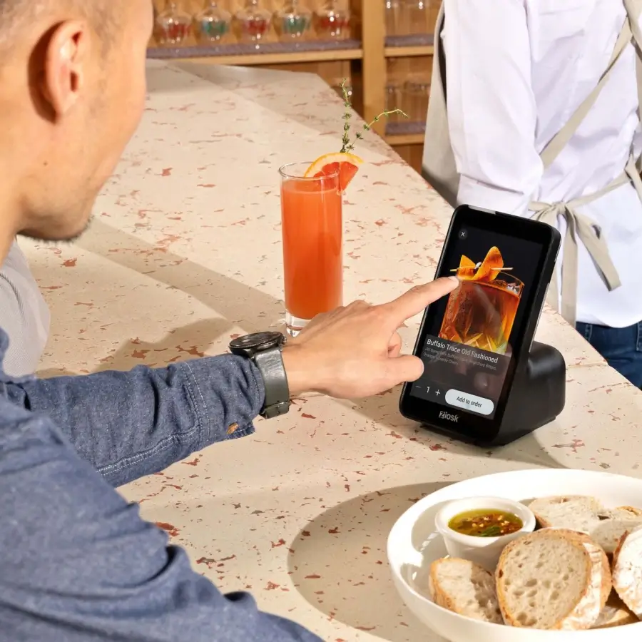 Person using a touchscreen kiosk to order a Buffalo Trace Old Fashioned cocktail, with a glass of orange drink and a plate of bread with dipping sauce on the table.