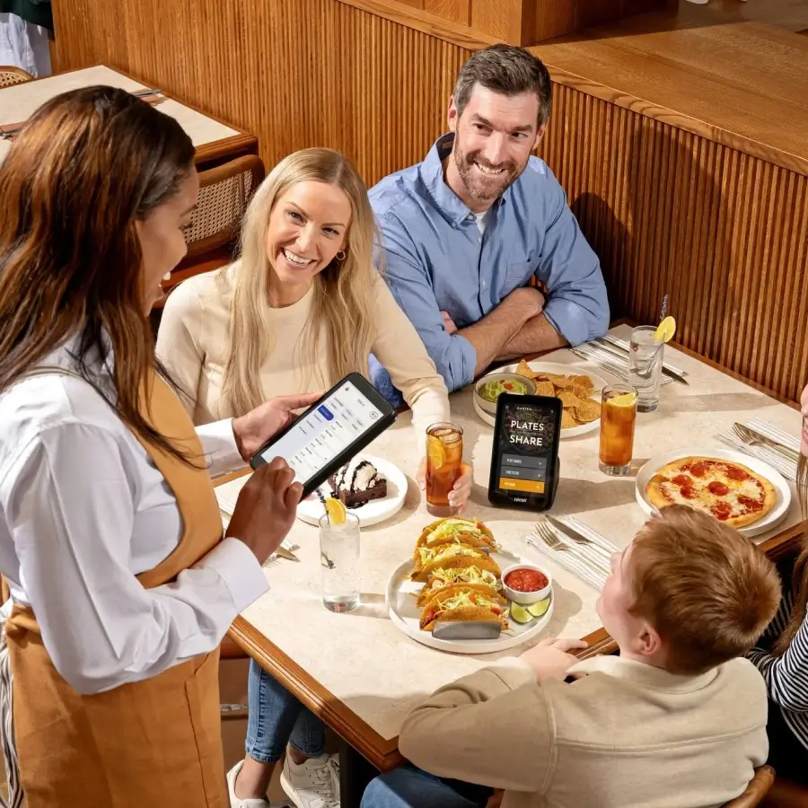 Waitress taking order from smiling family seated at a restaurant table with tacos, pizza, dessert, and drinks.
