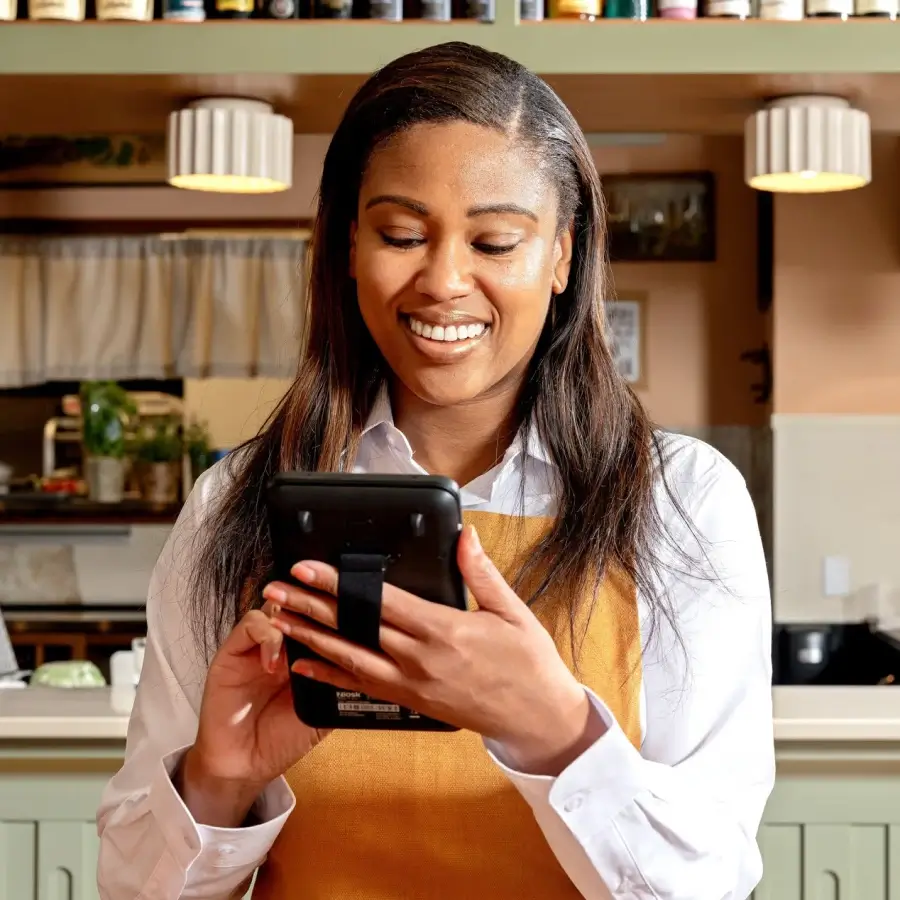 Smiling woman in a white shirt and brown apron holding and using a touchscreen device in a kitchen setting.