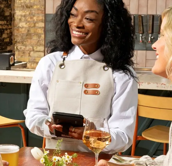 Smiling waitress in white apron holding a handheld device at a table with drinks and flowers.