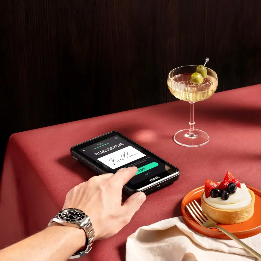 Hand with a wristwatch signing on a digital payment device on a red tablecloth next to a cocktail glass with olives and a small dessert with berries on an orange plate.