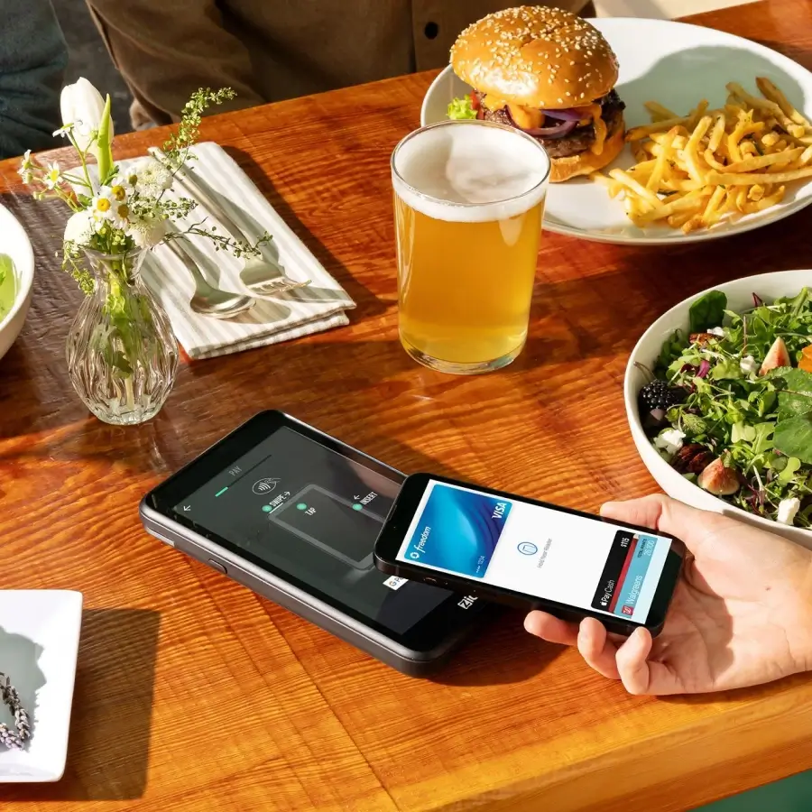 Person using smartphone with digital Visa card to make contactless payment on a card reader at a table with a burger, fries, salad, a glass of beer, and a vase with flowers.