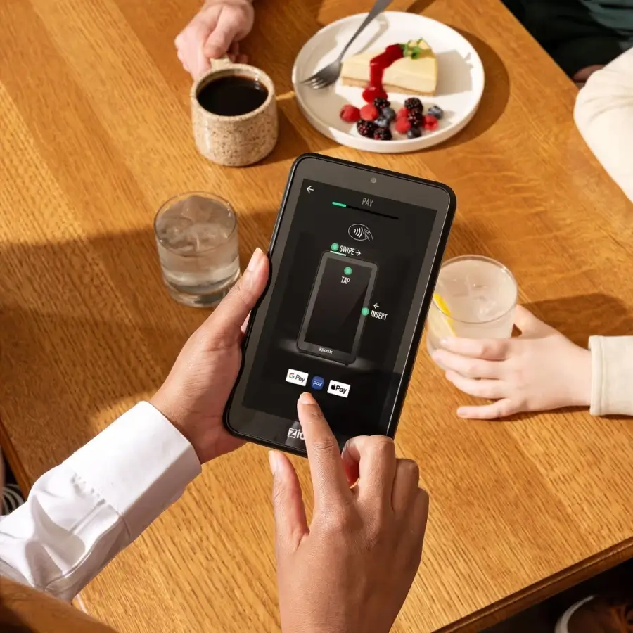 Person holding a digital payment device on a wooden table with coffee, dessert, and drinks.