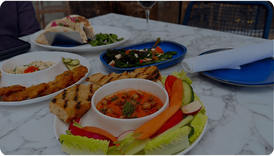 Table with plates of Mediterranean-style food including grilled bread, vegetable dip, fresh vegetable sticks, salad, pita wraps, and a glass of red wine.