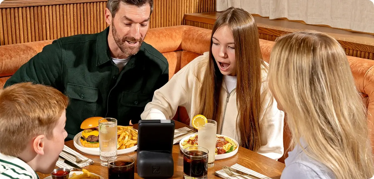 Family of four sitting at a wooden table with food and drinks, a girl excitedly looking at a device on the table.