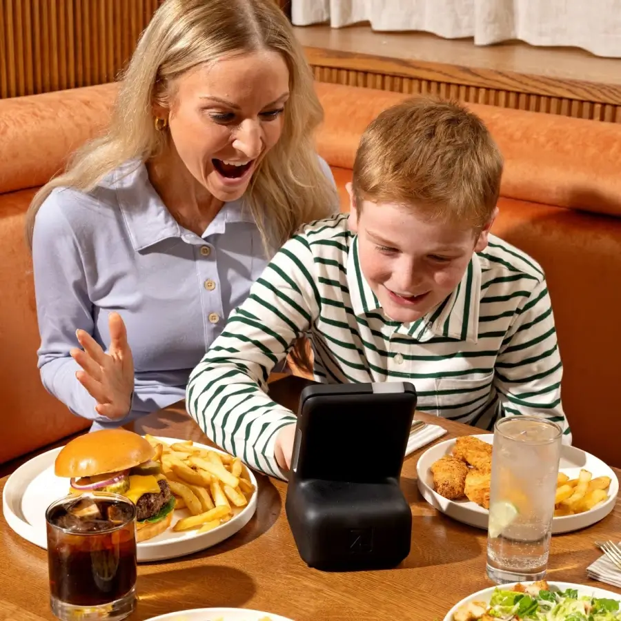 A woman smiling excitedly next to a boy who is focused on a handheld gaming device at a restaurant table with plates of fries, a burger, chicken nuggets, and drinks.