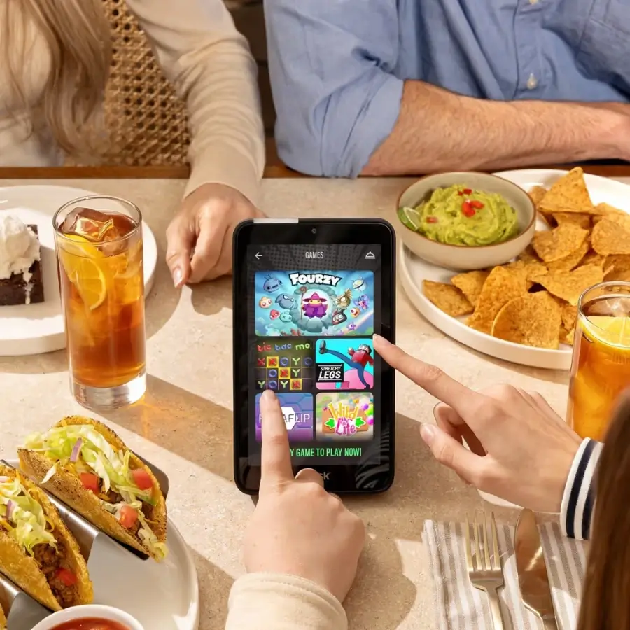 Two people seated at a table with tacos, iced tea, chips and guacamole, and a dessert, while they select a game on a tablet screen.