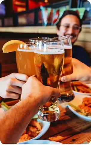 Three people clinking beer glasses together over a wooden table with plates of food in a casual dining setting.