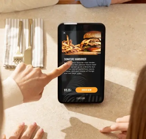 Person using digital menu on a tablet showing a signature hamburger with fries and an order button at a restaurant table with utensils.