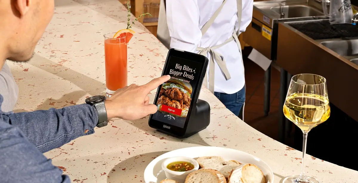 Person touching a digital kiosk screen on a counter displaying a food deal advertisement next to a glass of orange drink and a plate of bread.