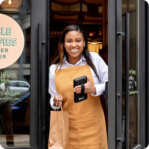 Smiling woman in a tan apron holding a tablet and a brown paper bag stands in a doorway of a store or cafe.