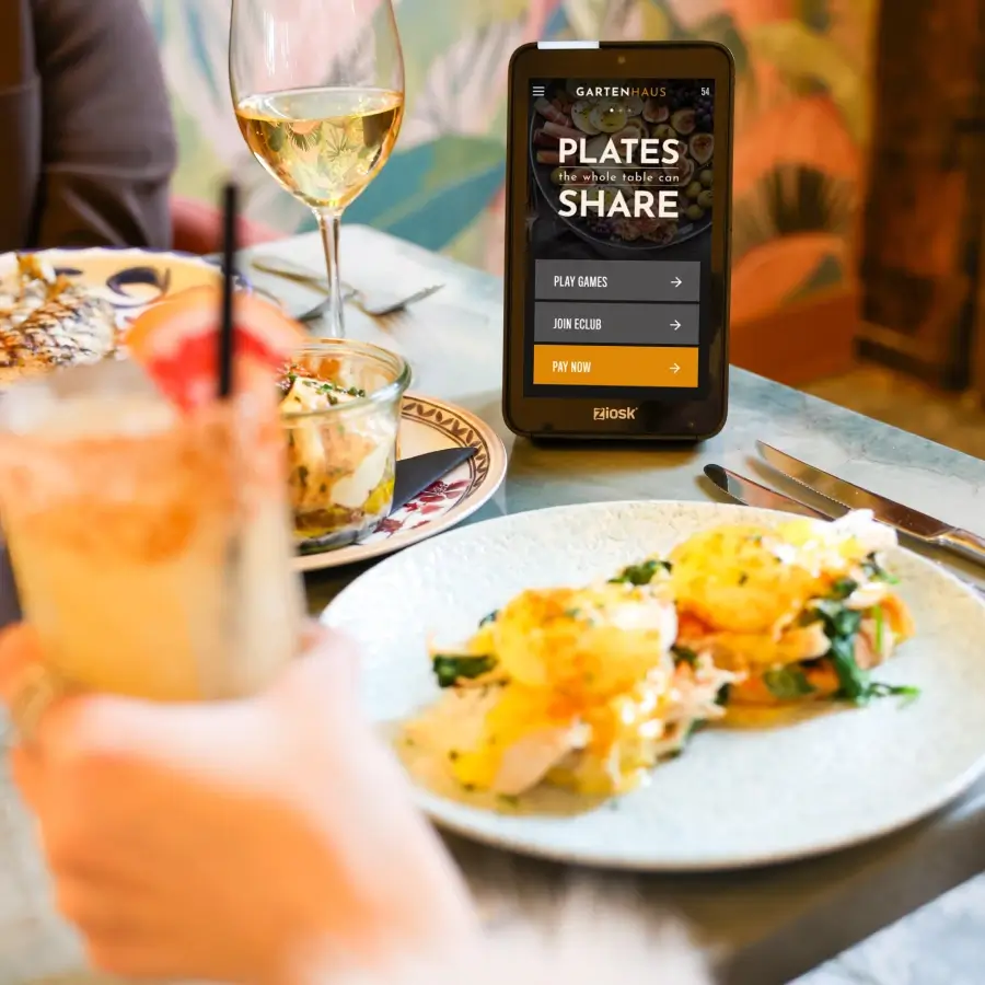 Restaurant table with plated food, a glass of white wine, and a Ziosk device showing options to play games, join eClub, or pay now.