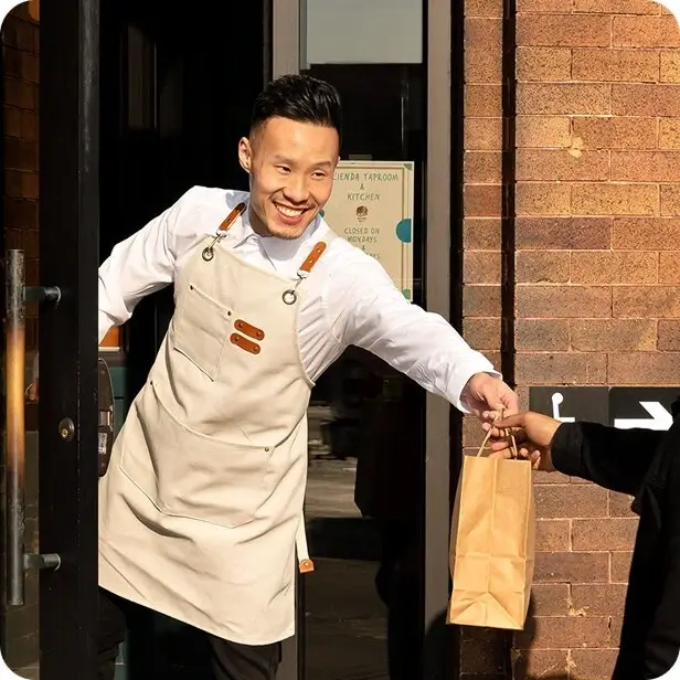 Smiling man in beige apron handing a brown paper bag to a customer outside a brick building.