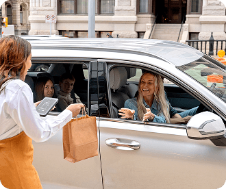 Woman handing a paper bag to a smiling female driver in a silver car with two passengers in the back seat on a city street.