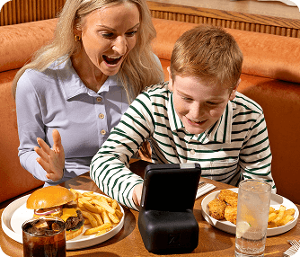 Woman and boy excitedly looking at a black electronic device on a restaurant table with plates of fries, a burger, fried food, and drinks.