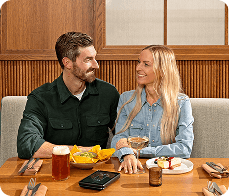 Smiling man and woman sitting at a restaurant table with food and drinks, looking at each other.