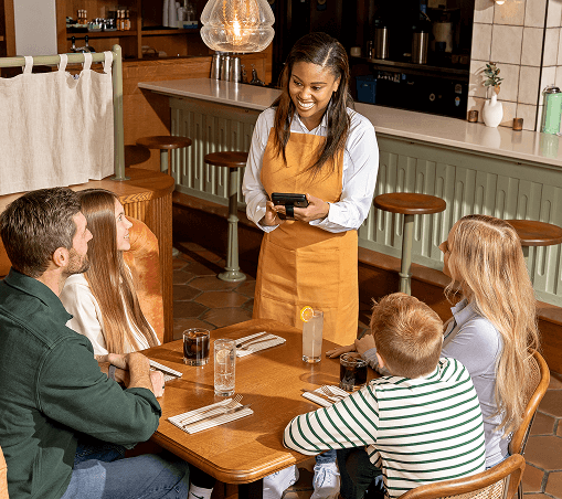 Smiling waitress taking order from family of four seated at a wooden table in a restaurant.