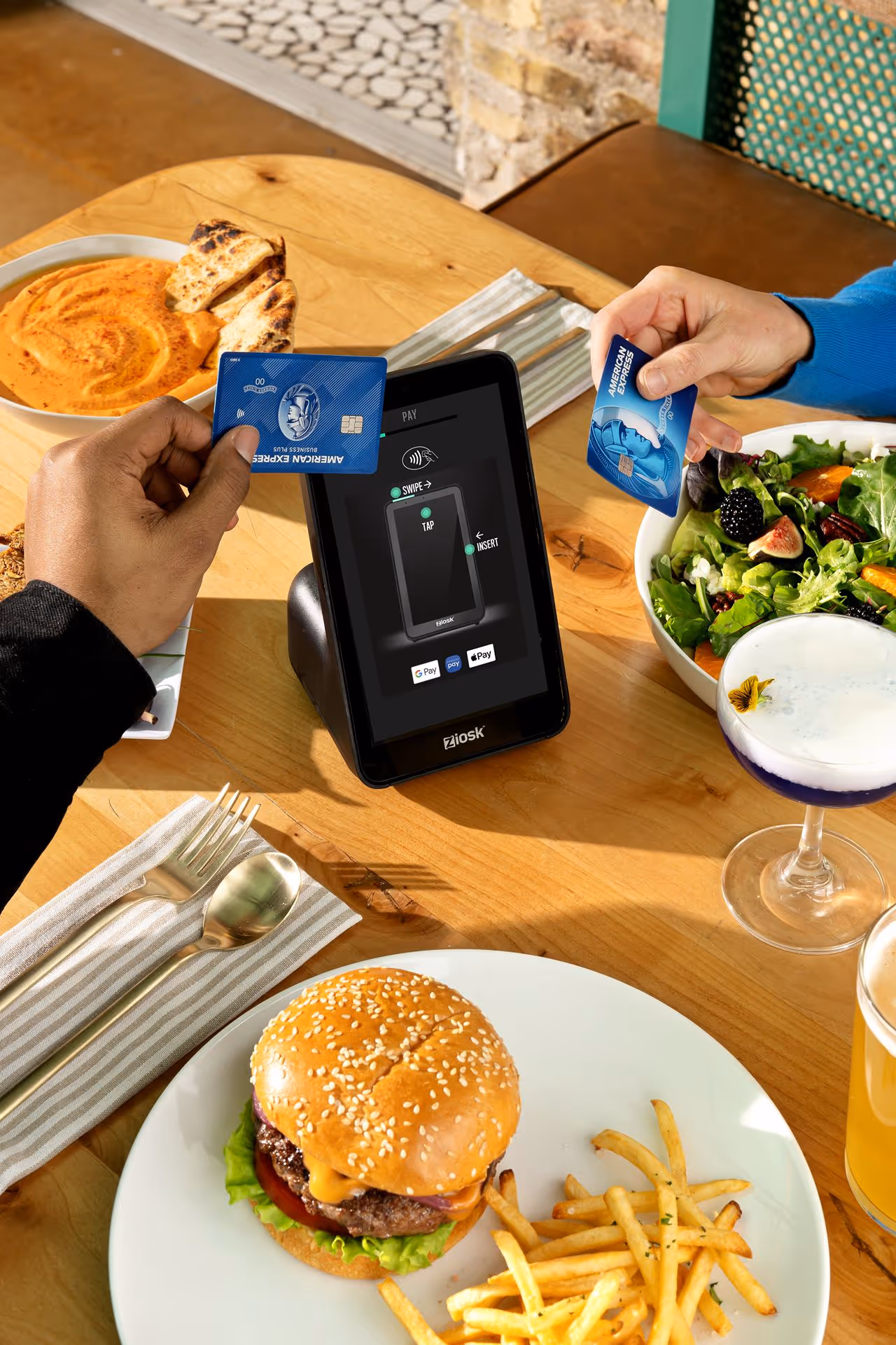 Two people holding American Express cards near a Ziosk payment terminal on a table with a burger, fries, salad, dipping sauce, and drinks.
