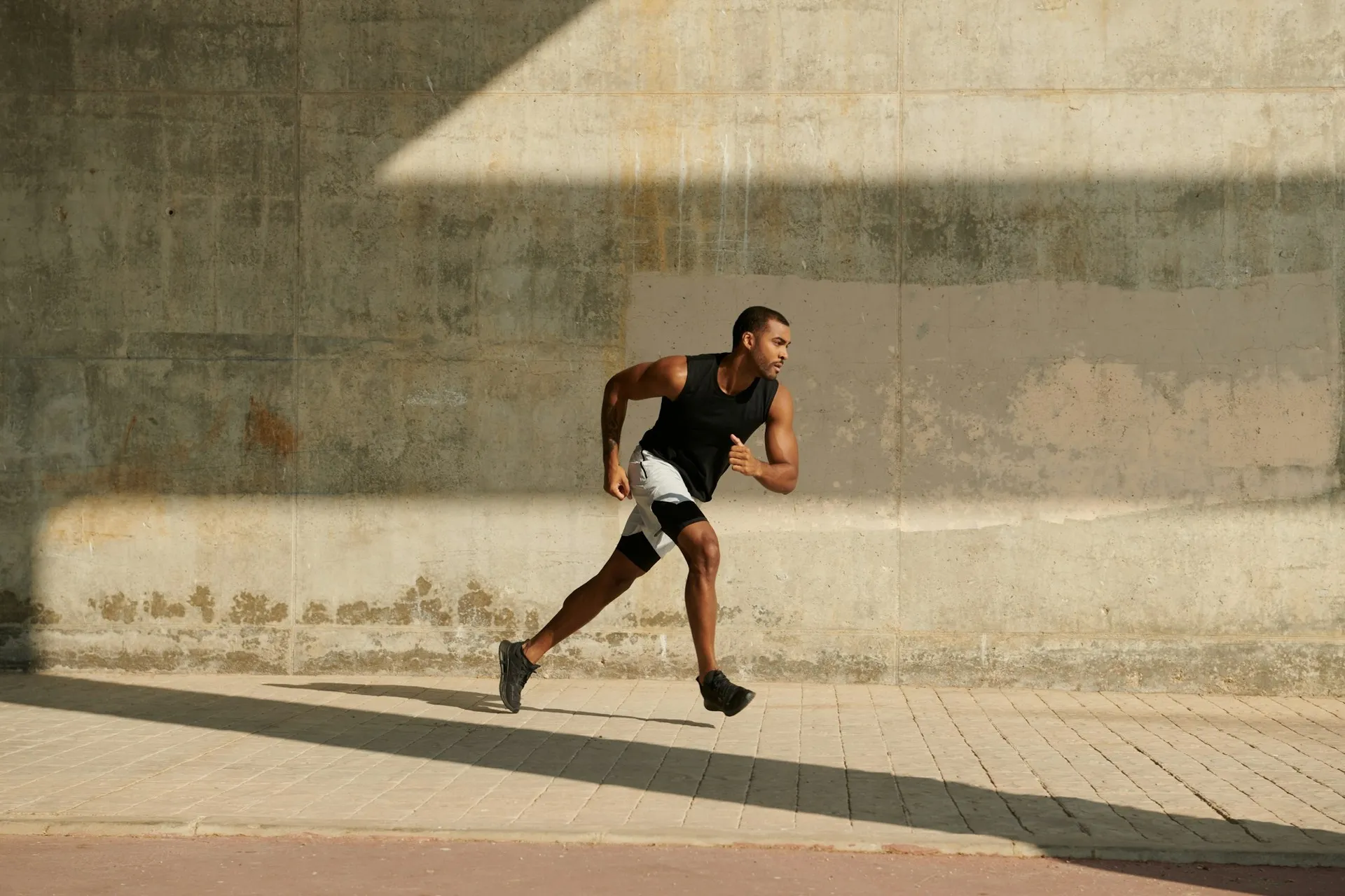 A man in a black singlet and white shorts running to an urban backdrop of a concrete wall and sandstone footpath.