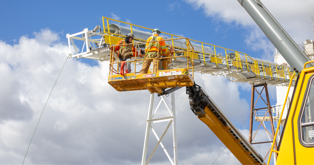 Two construction workers in safety gear standing on a yellow aerial lift platform against a blue sky with clouds.