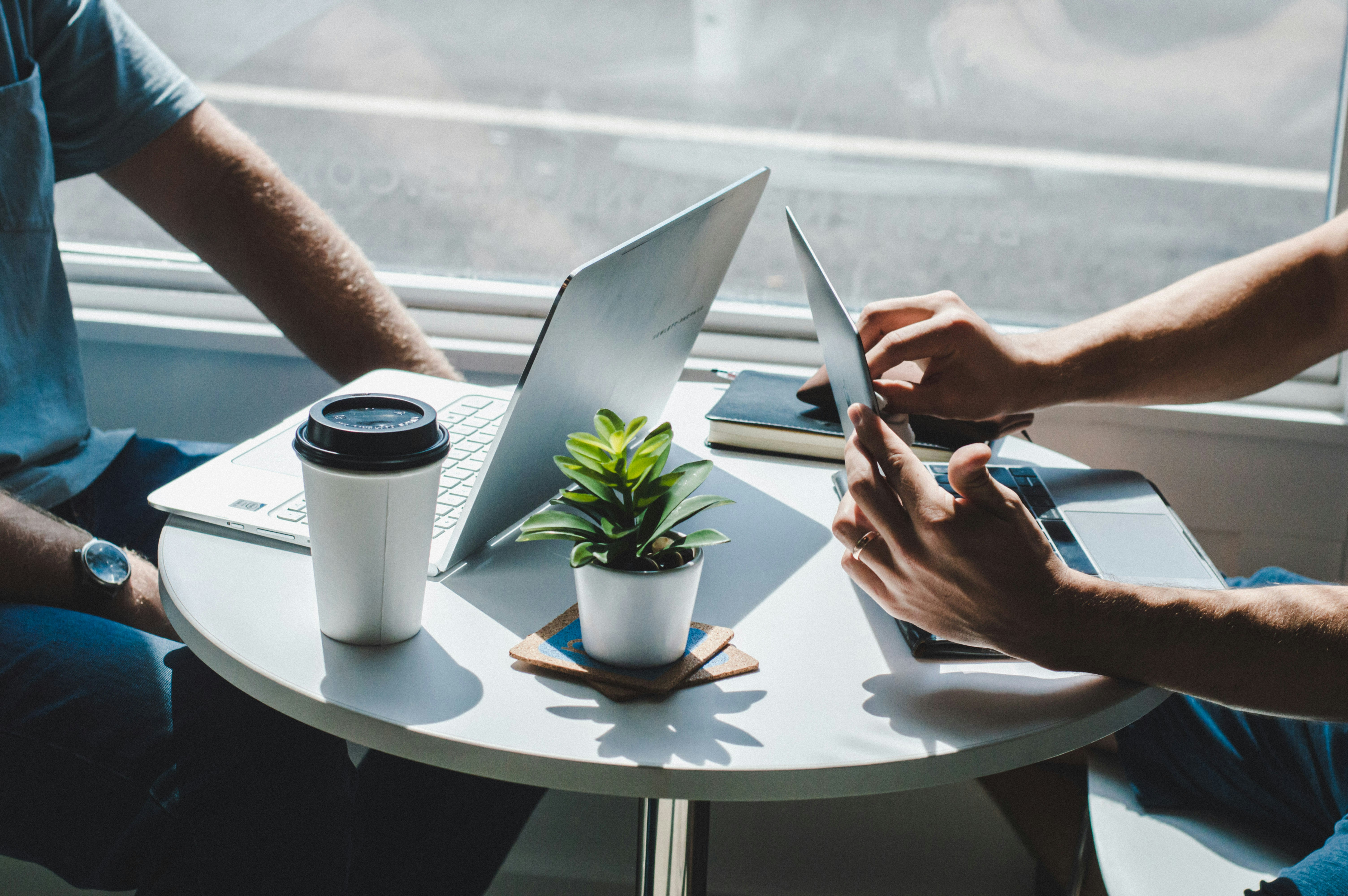 Two people sitting at a round table with laptops, a coffee cup, a small potted plant, and a notebook near a window.