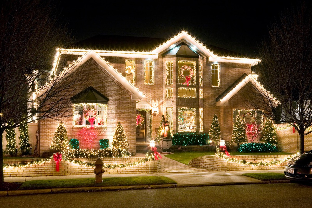 Two-story brick house decorated with white Christmas lights, wreaths with red bows, and a snowman figure on the front lawn at night.