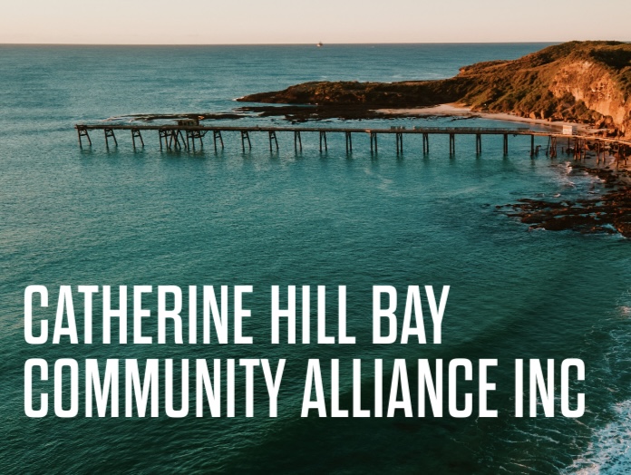 Wooden pier extending over blue-green ocean water toward a rocky, shrub-covered coastline under a clear sky at sunset.