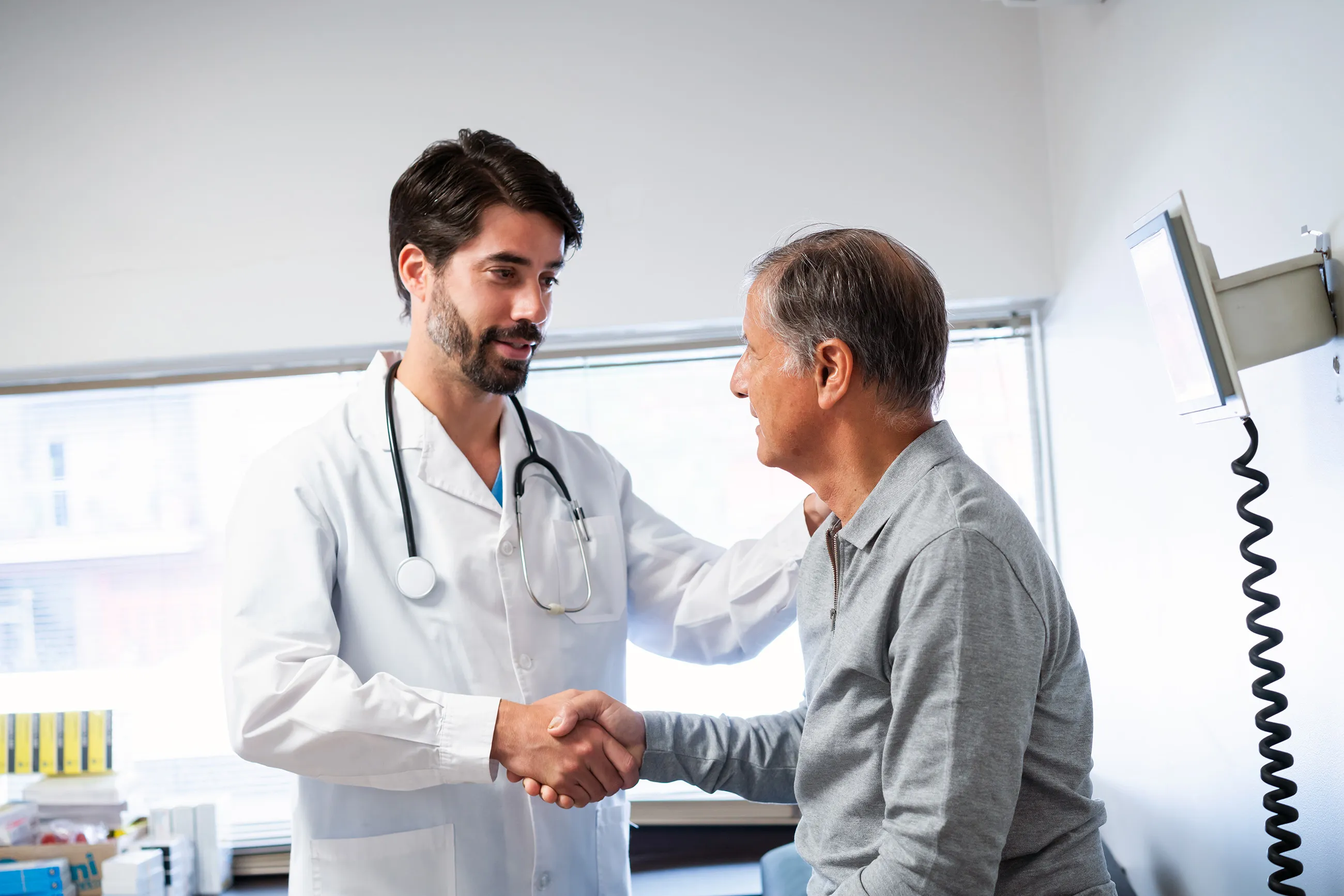Male doctor in white coat shaking hands and talking with older male patient in clinic room.