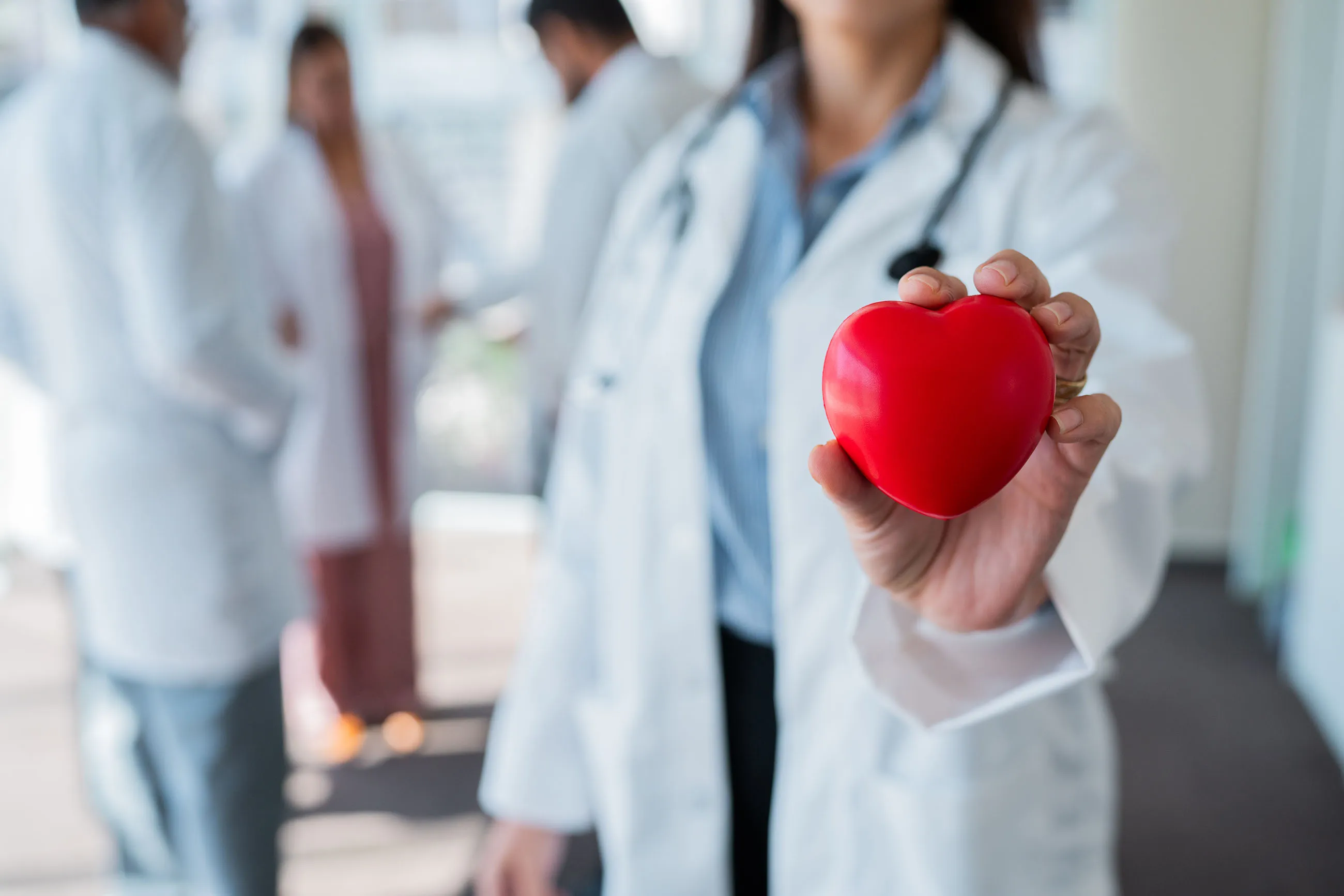 Medical professional holding a red heart-shaped object with blurred healthcare workers in the background.