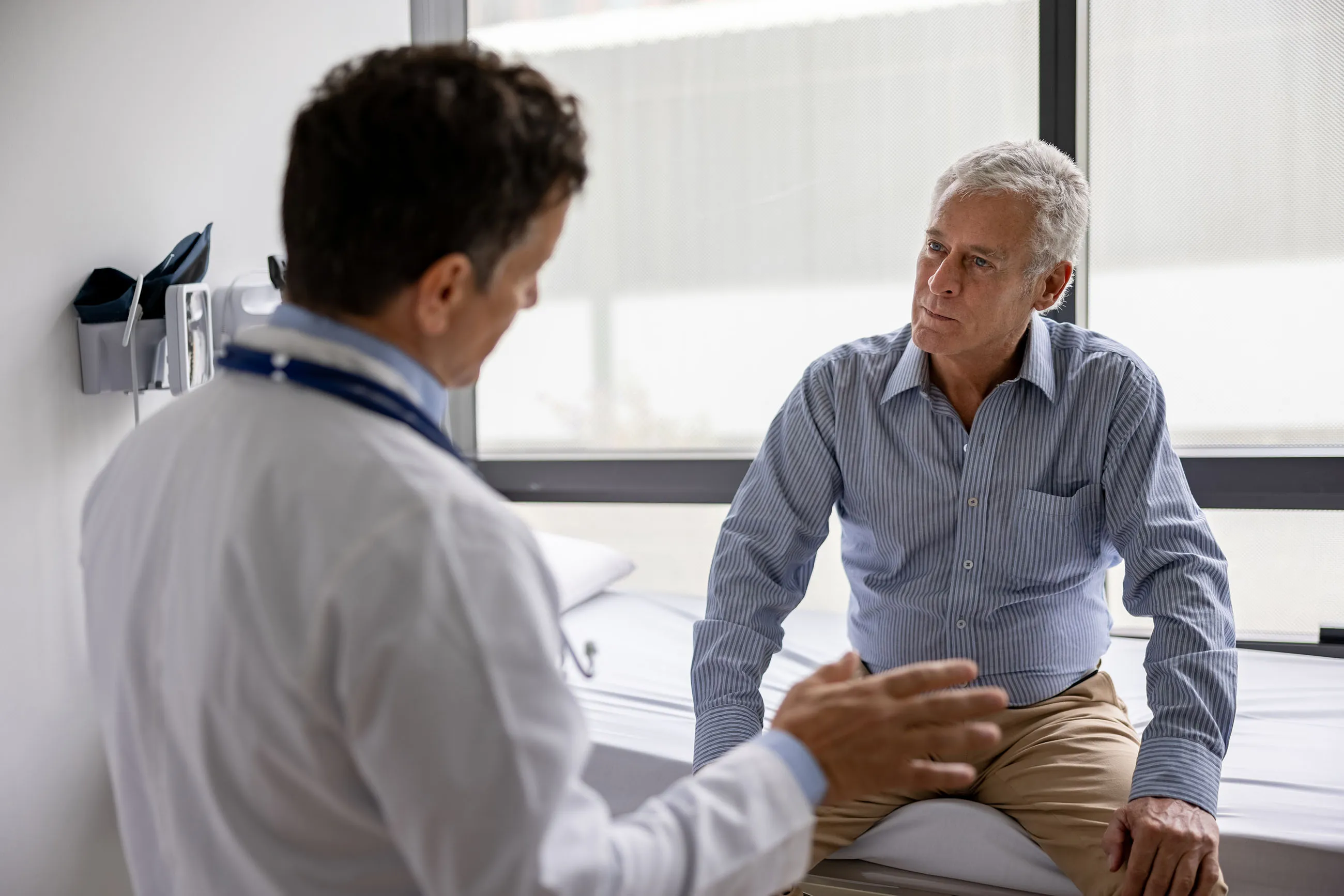 Male doctor in a white coat talking to an older male patient sitting on a hospital bed.