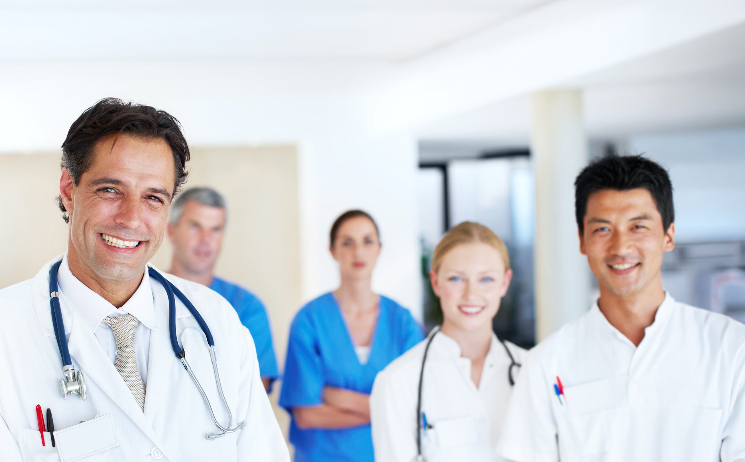 Smiling diverse medical team of five doctors and nurses in a bright hospital corridor.
