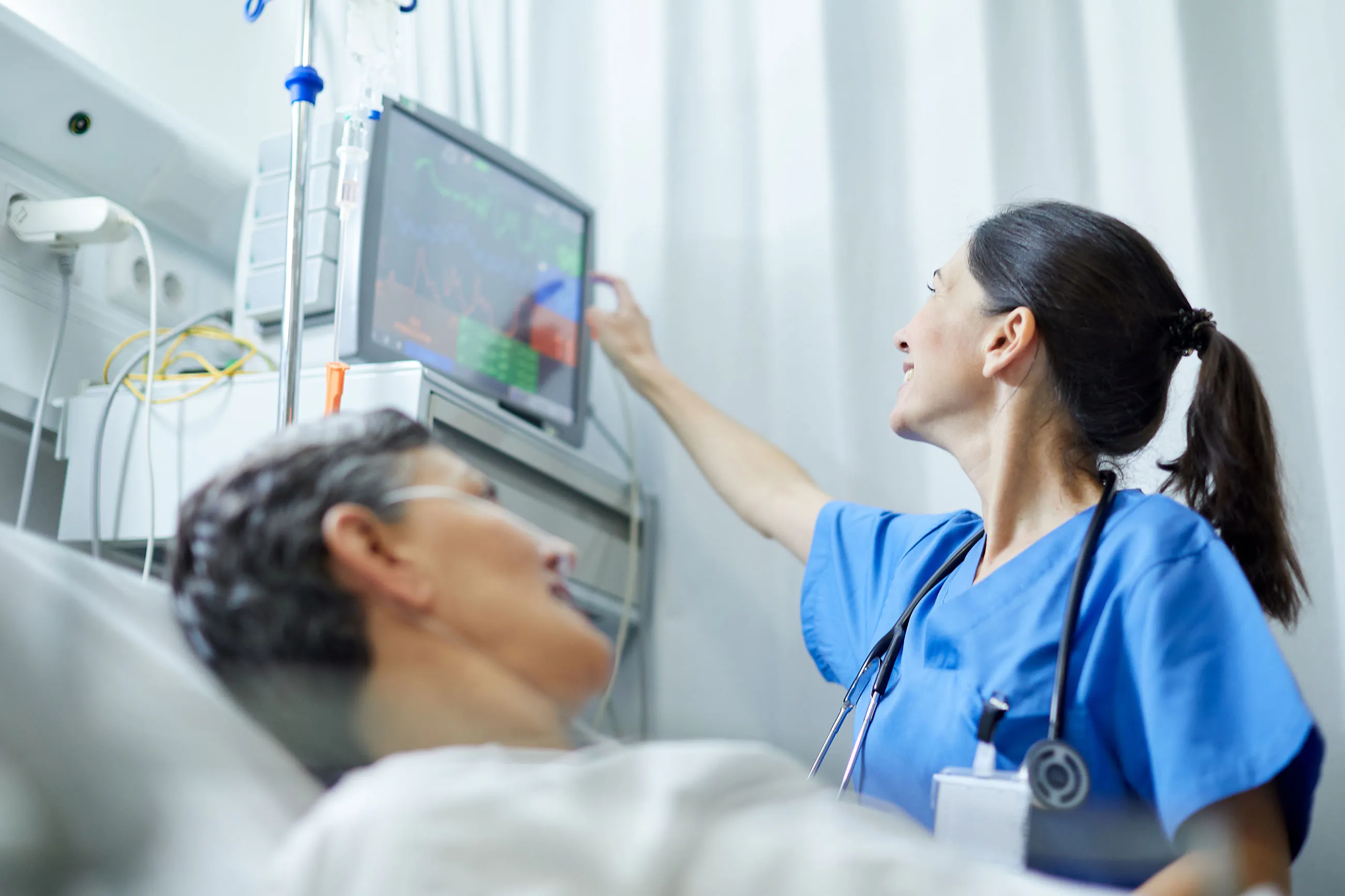 Nurse in blue scrubs monitoring a patient's vital signs on a screen in a hospital room.
