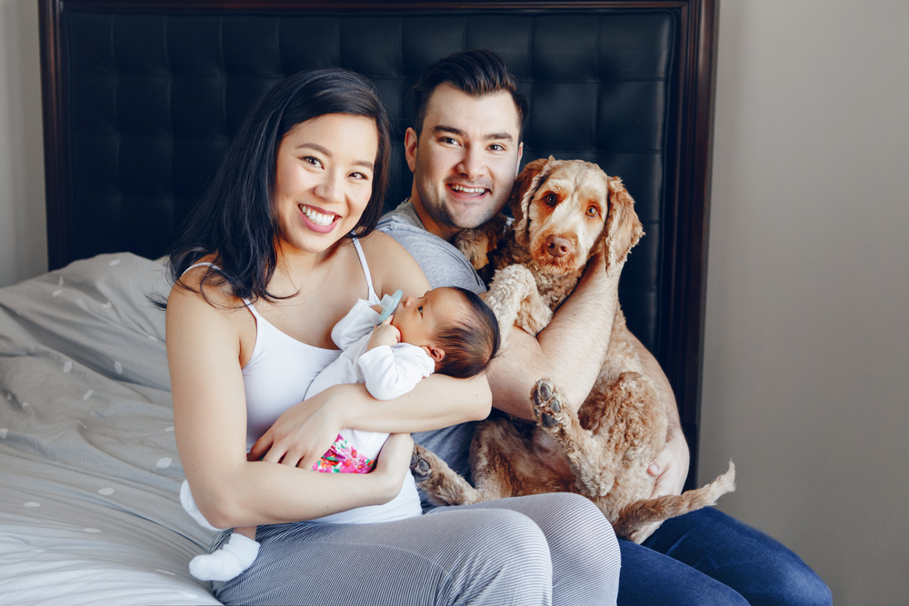 A woman sitting holds a newborn baby. A man sits beside her holding a dog.