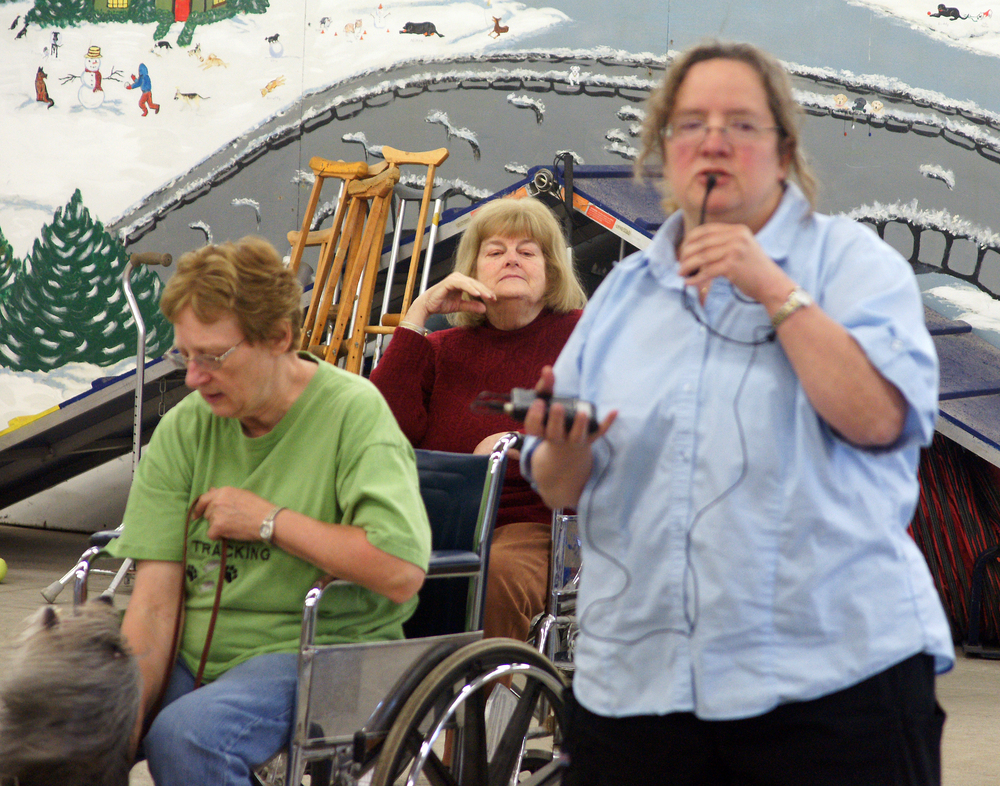 At a dog school, a woman holds a microphone as if speaking toward the camera to an unseen audience, while two other women sit in wheelchairs beside her.