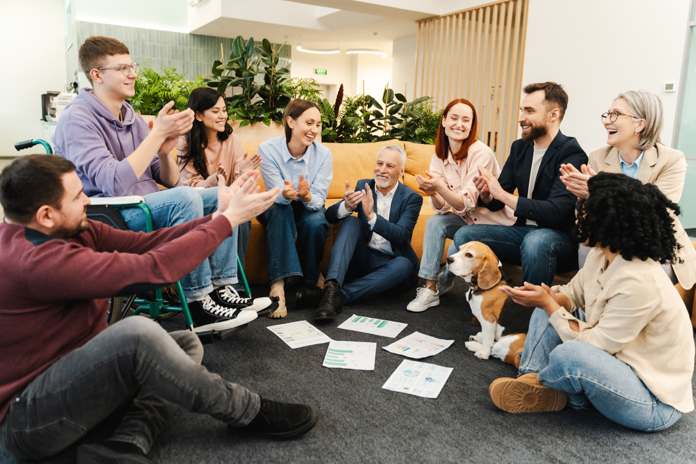 Nine young adults sit in a semi-circle in a relaxed environment clapping as if celebrating an event. A small dog sits beside one of the people.
