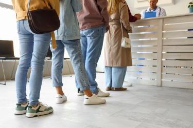 Patients stand in line at a clinic reception desk while a staff member assists them from behind the counter.