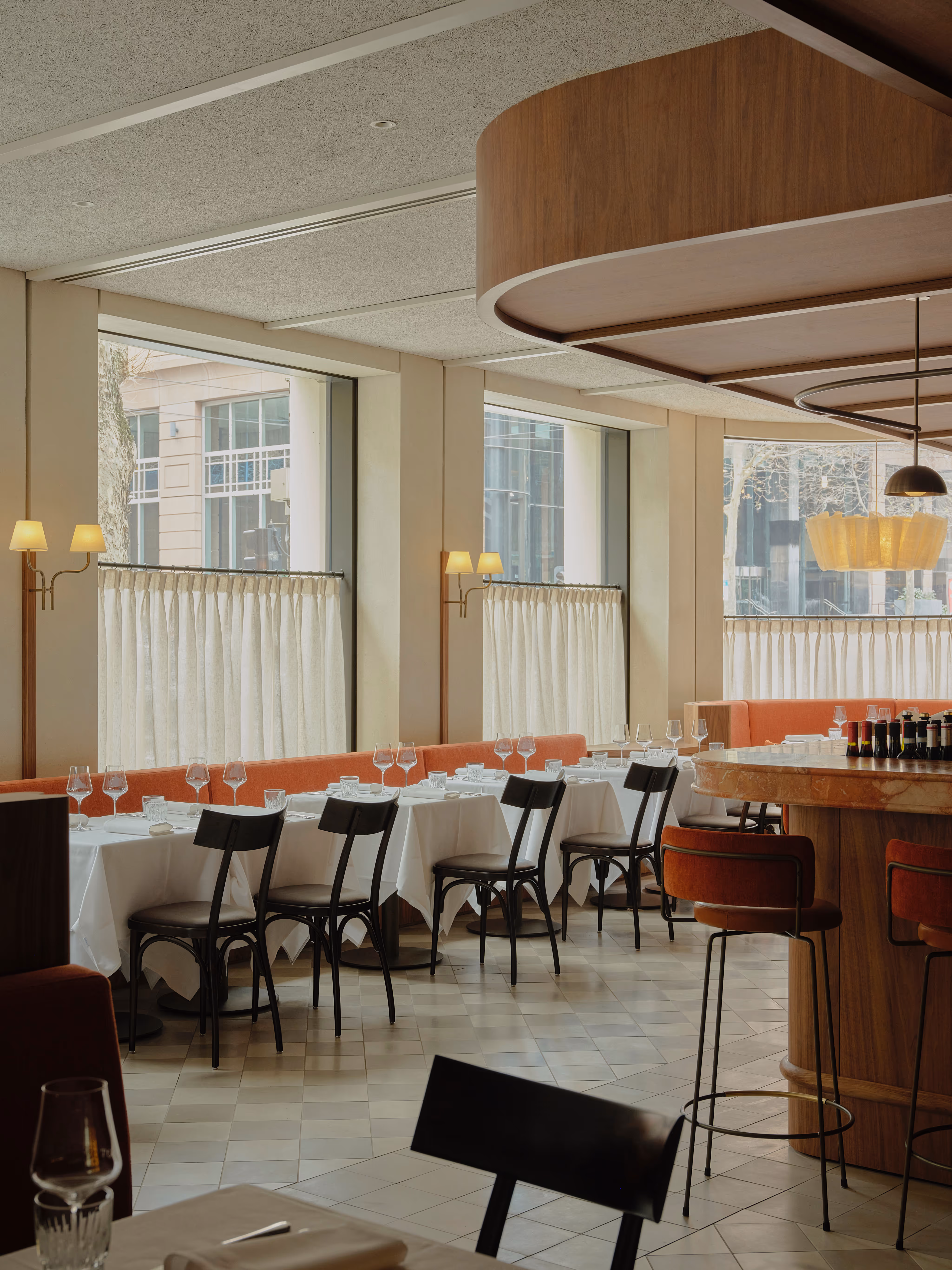 Modern restaurant interior with large windows, white tablecloth-covered tables, black chairs, and an orange booth seating alongside a circular wooden bar.