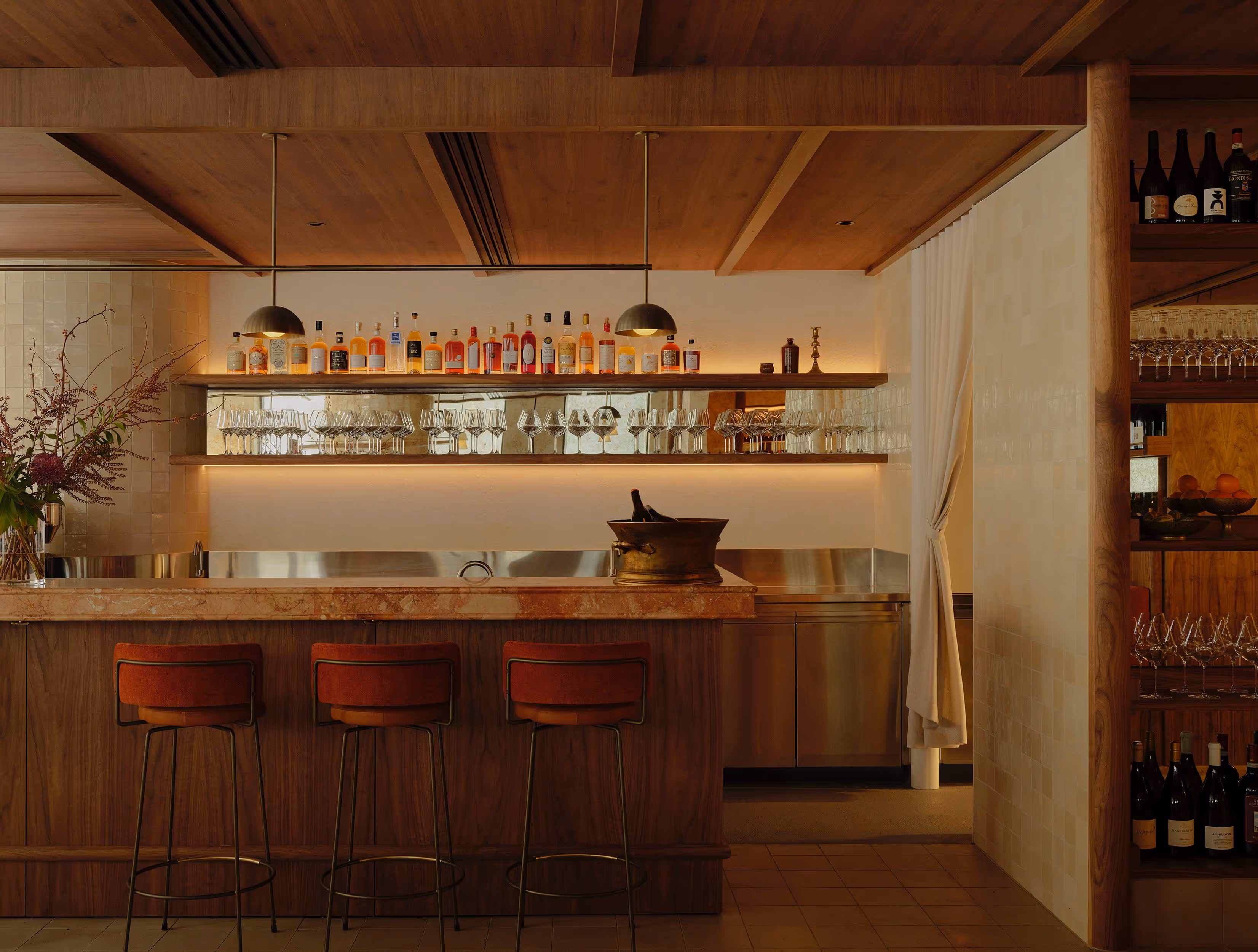 Cozy wooden bar interior with three red cushioned bar stools, illuminated liquor bottles and wine glasses on shelves, and a floral arrangement in a vase.