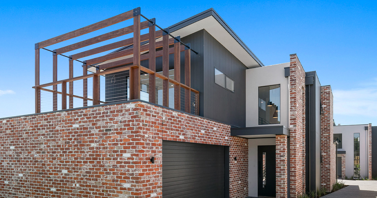 Modern two-story house with red brick facade, dark garage door, and wooden pergola on the upper balcony under a clear blue sky.