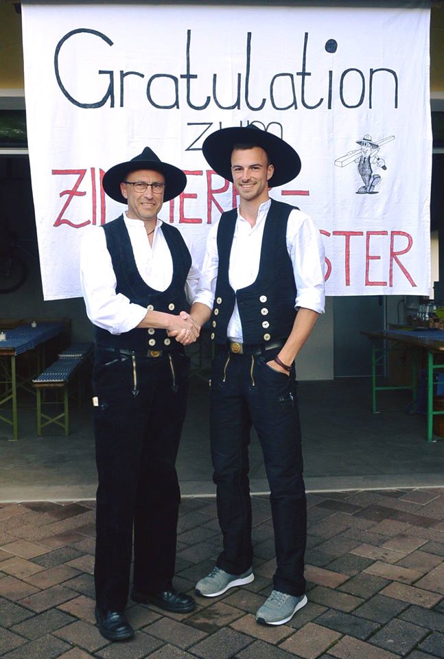 Two men wearing traditional carpenter outfits with hats shaking hands in front of a congratulatory banner.