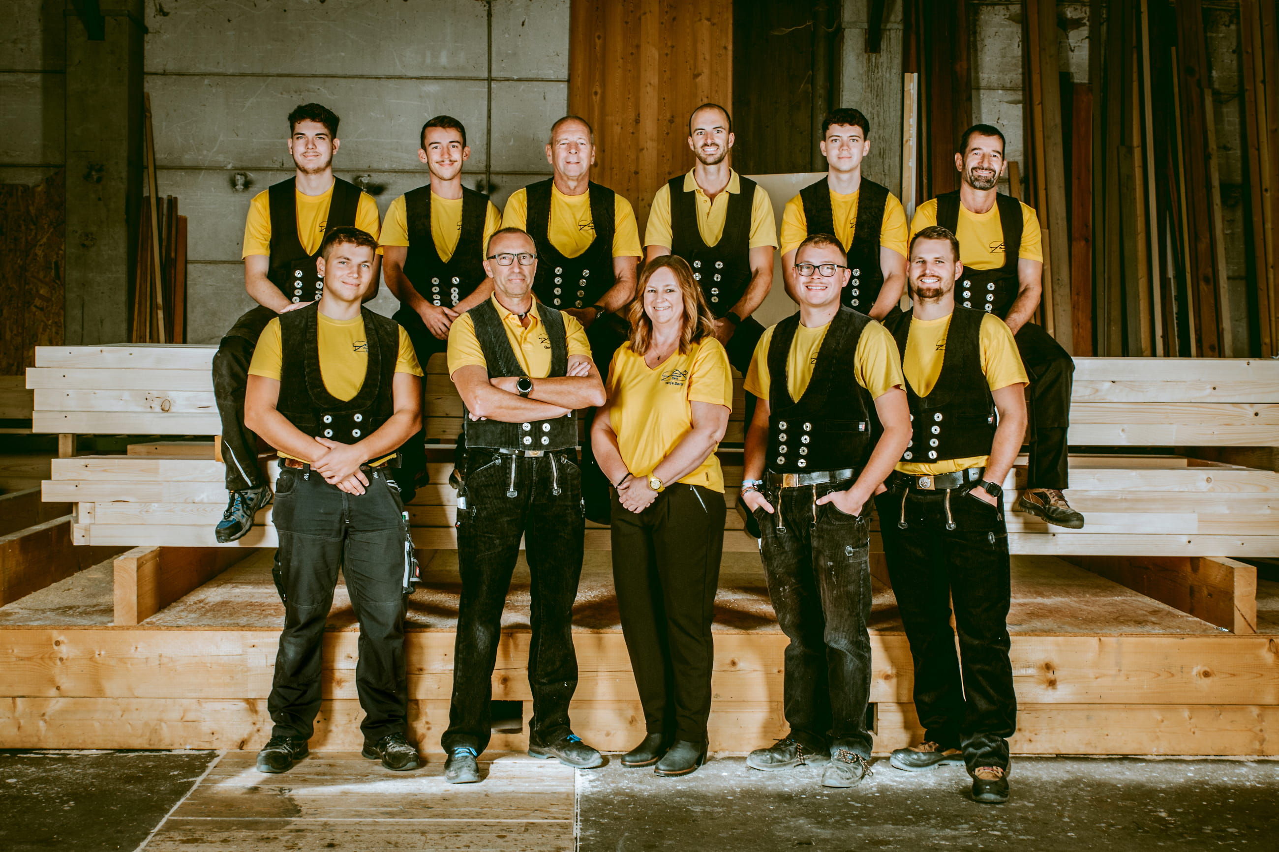 Group of ten workers in yellow shirts and black vests posing inside a woodworking shop.