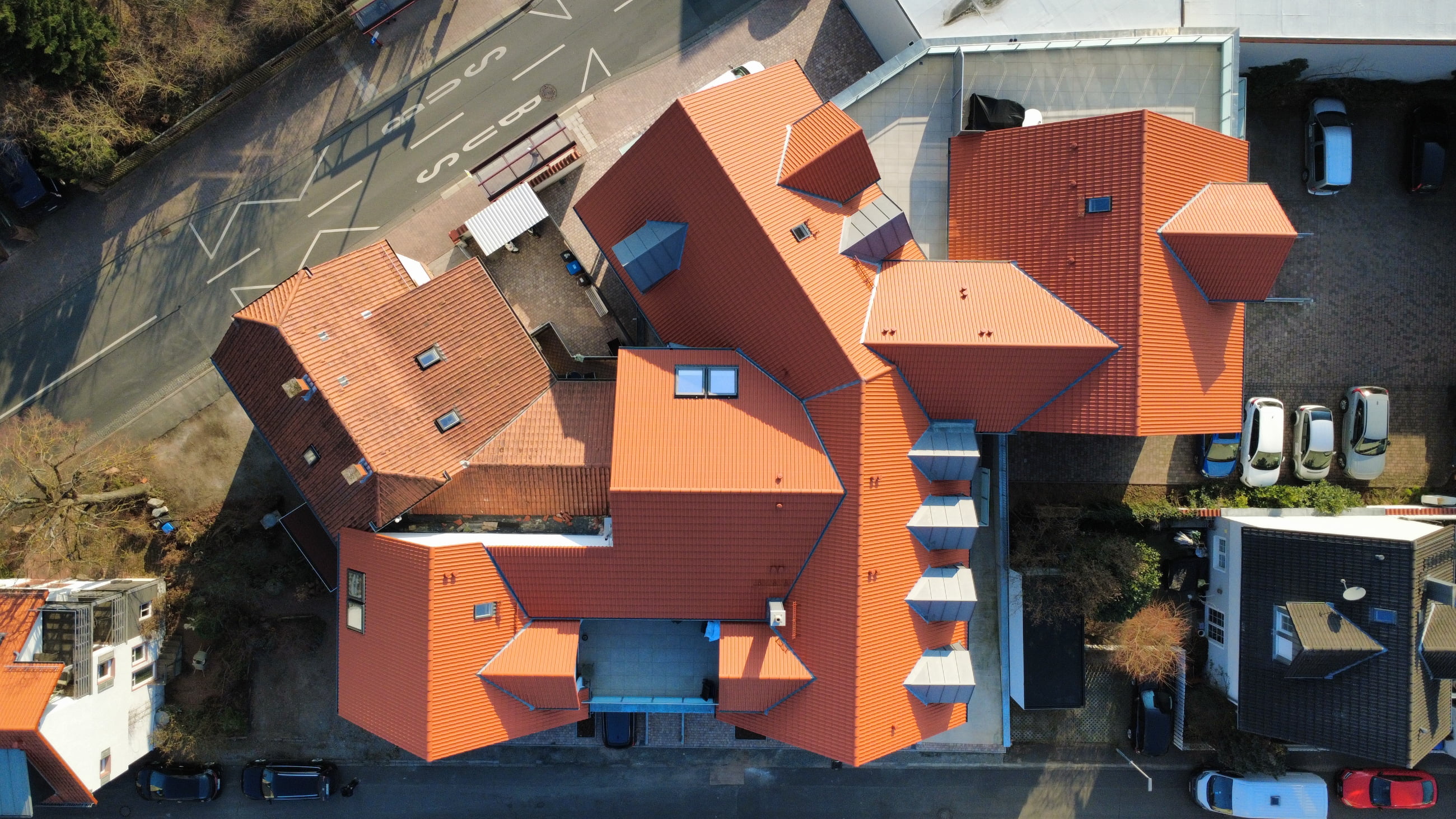 Aerial view of a large building with complex red-tiled roofs and surrounding parked cars.