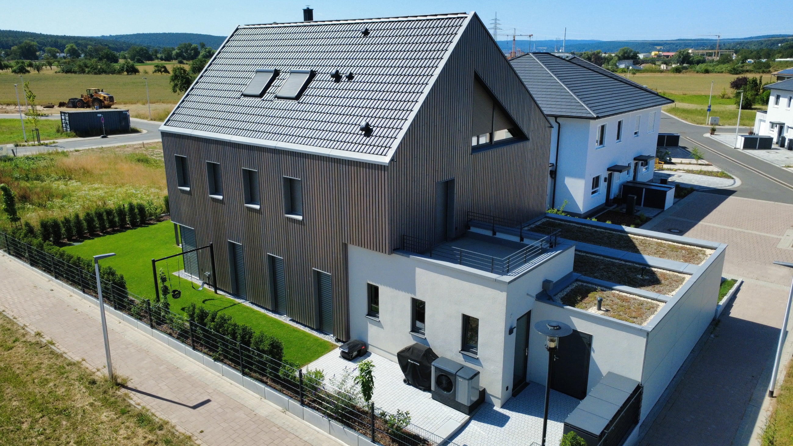 Modern two-story house with a dark tiled roof and wooden facade, surrounded by a well-maintained green lawn and hedges.