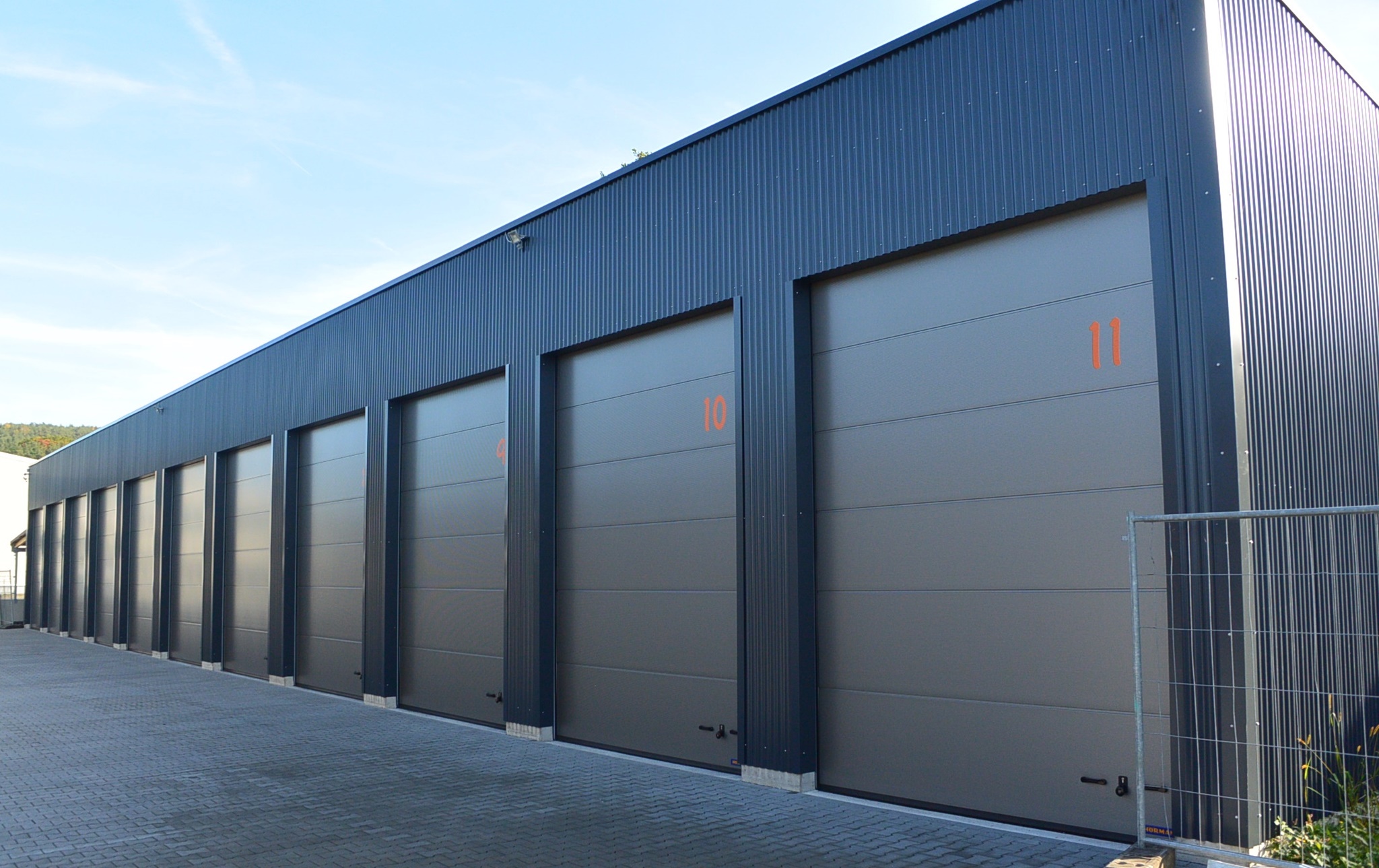 Row of dark gray industrial garage doors numbered from 8 to 11 on a modern warehouse building under a blue sky.