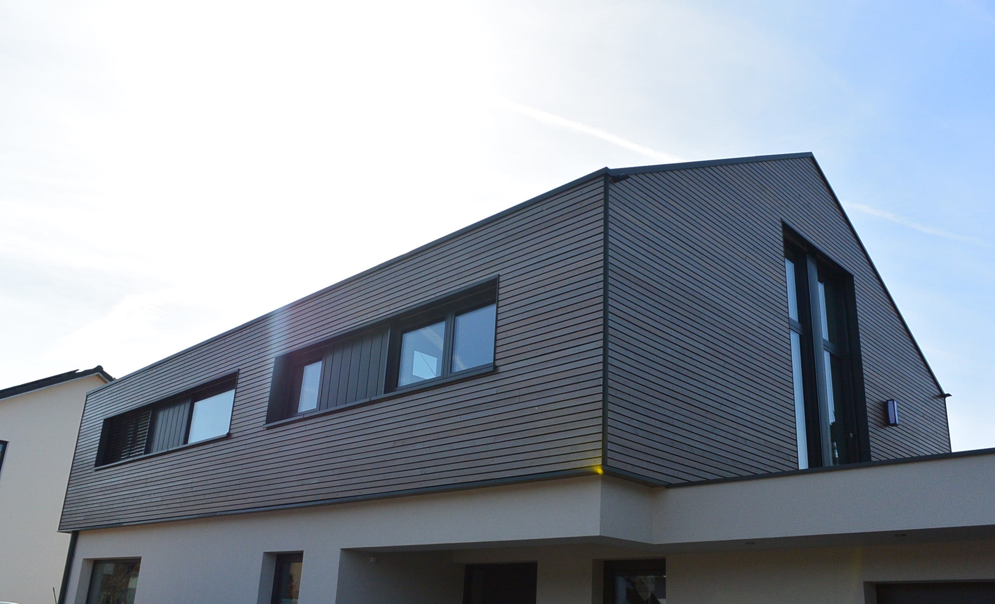 Modern two-story house with dark horizontal wooden siding on the upper floor and large windows in bright daylight.