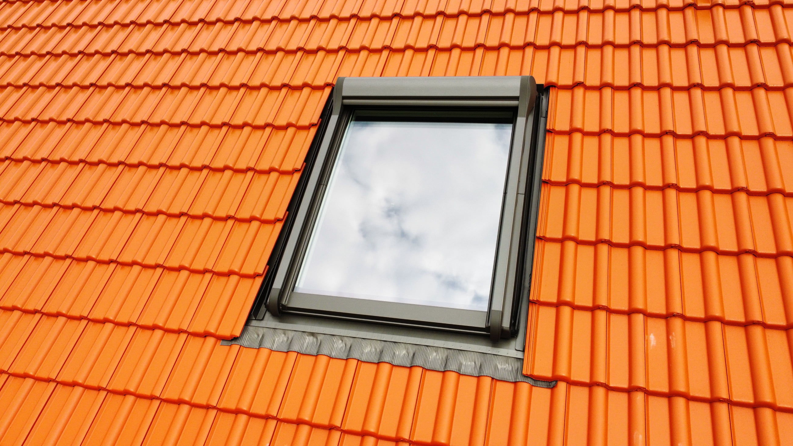 Gray skylight window reflecting clouds surrounded by bright orange roof tiles.