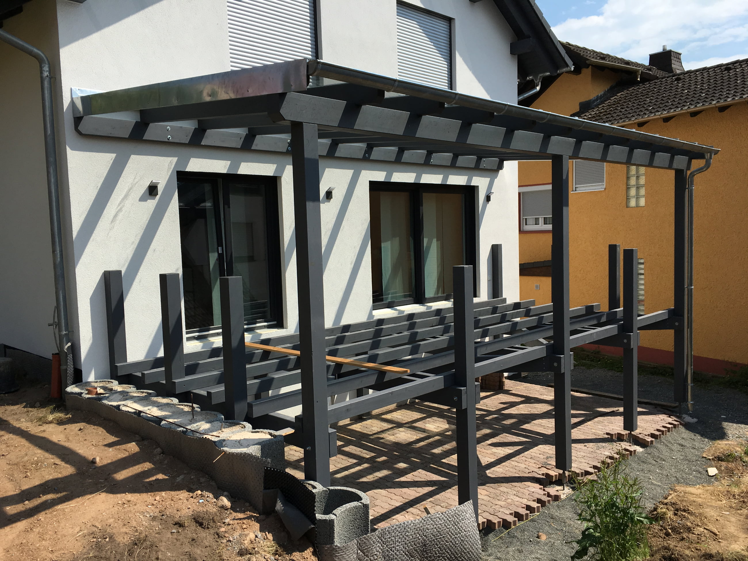 Partially constructed wooden patio frame attached to the back of a white house with closed windows and a paved ground area.
