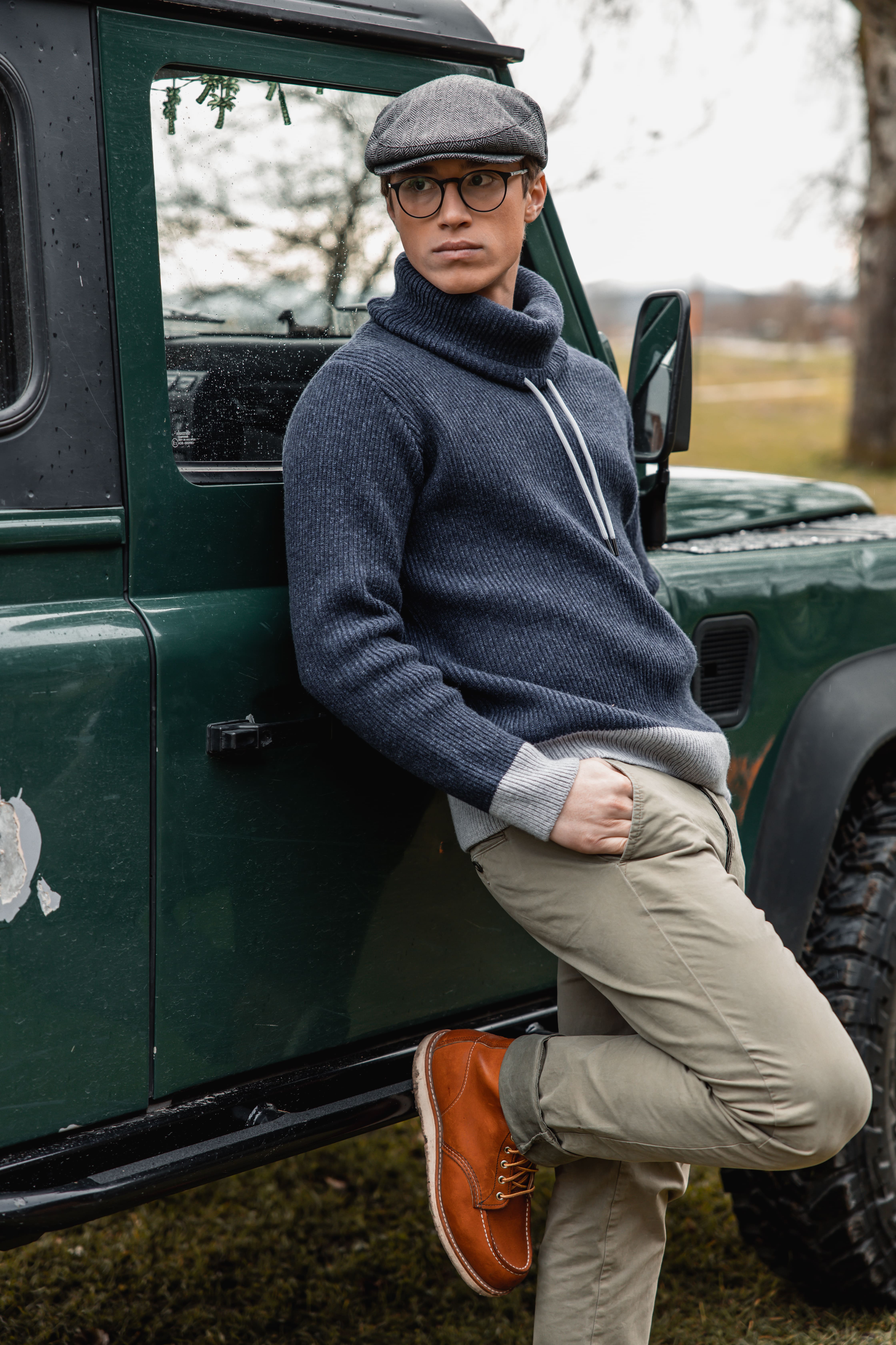 Young man in a gray flat cap, glasses, navy sweater, beige pants, and brown boots leaning against a green vehicle outdoors.