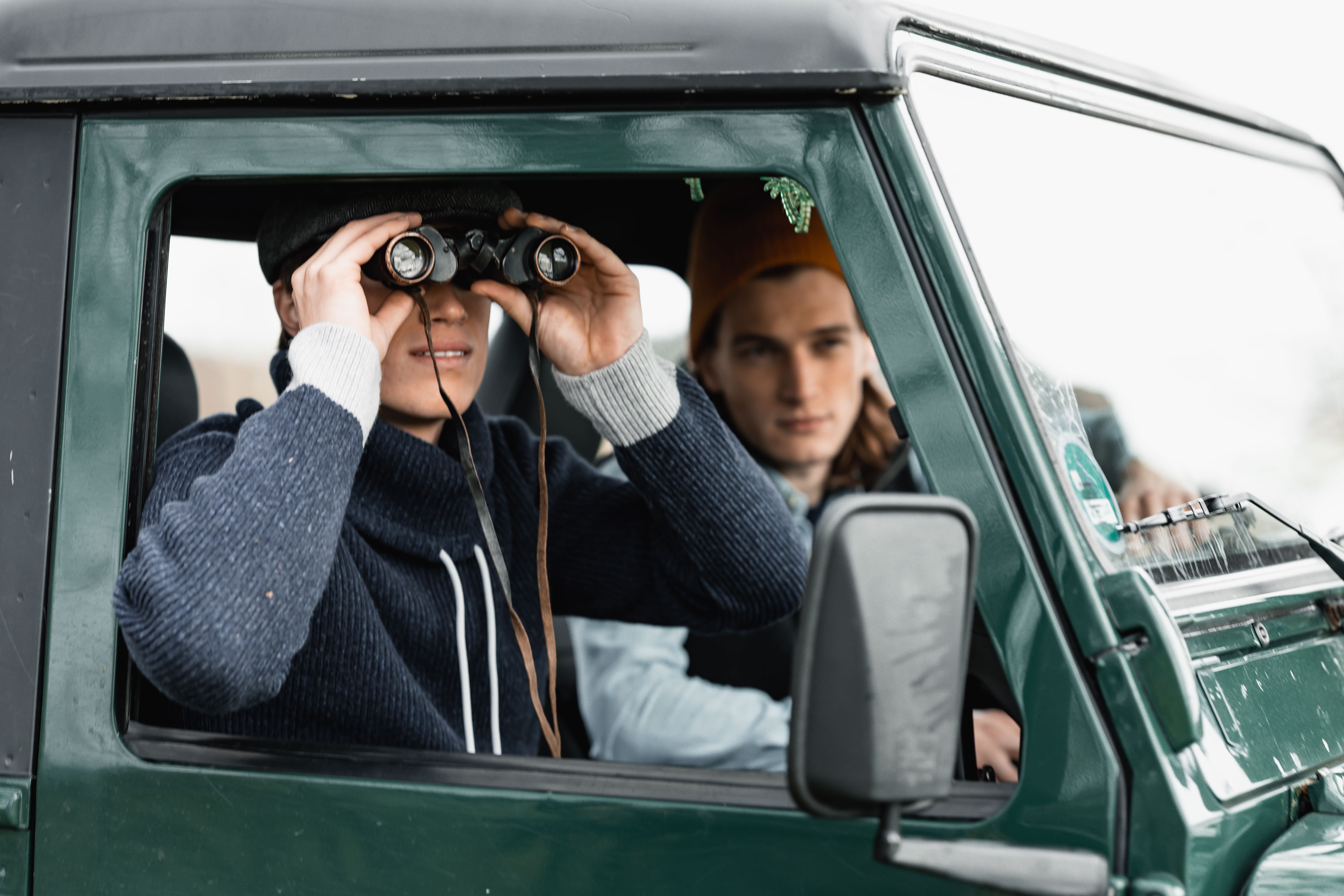 Two young men sitting inside a green vehicle, one looking through binoculars while the other looks forward.
