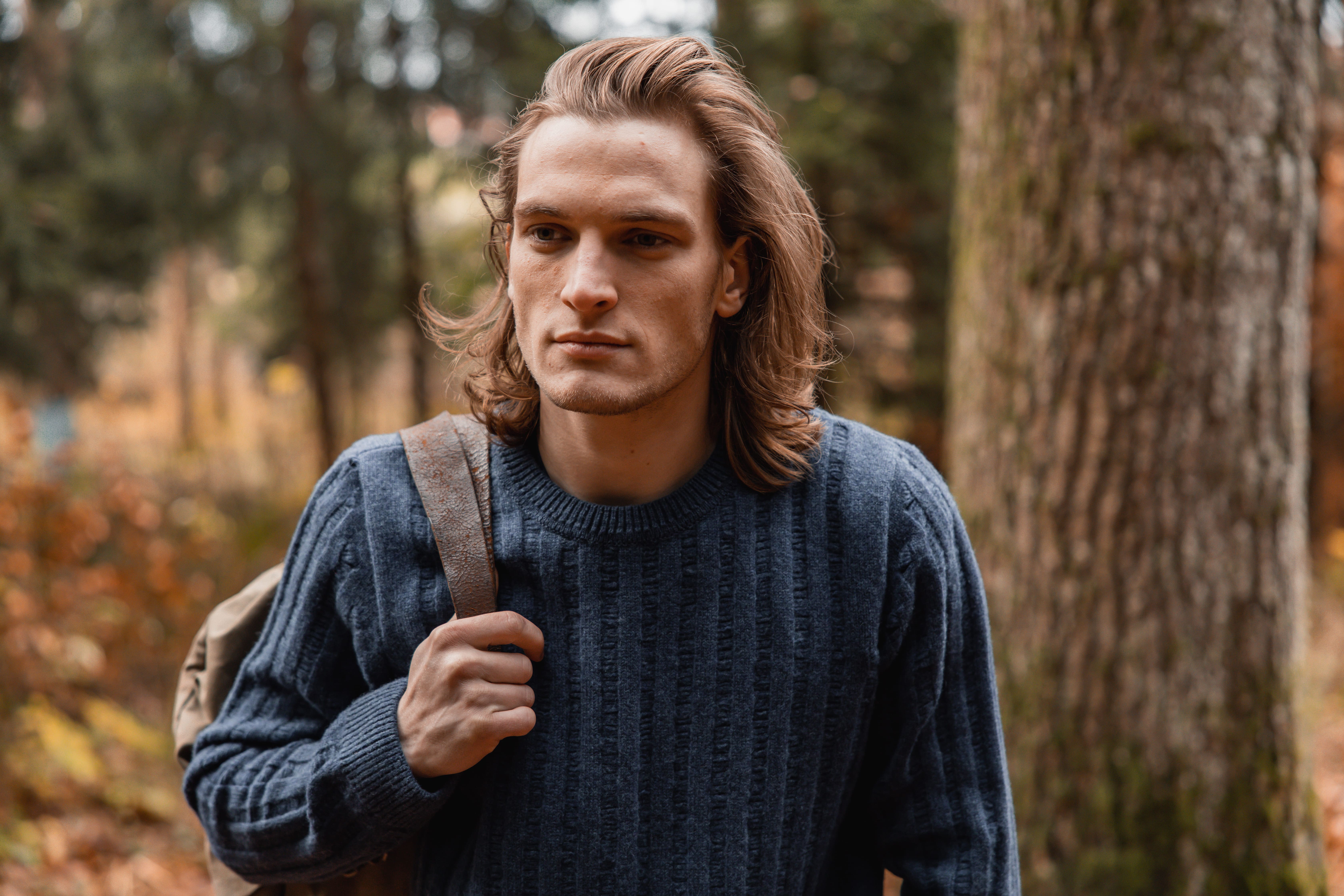 Young man with shoulder-length hair wearing a blue sweater and carrying a backpack in a forest.
